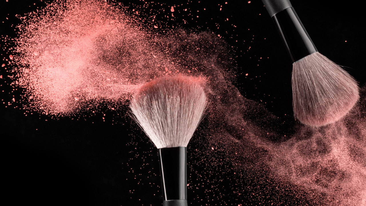 A makeup brush releasing a cloud of blush powder against black.